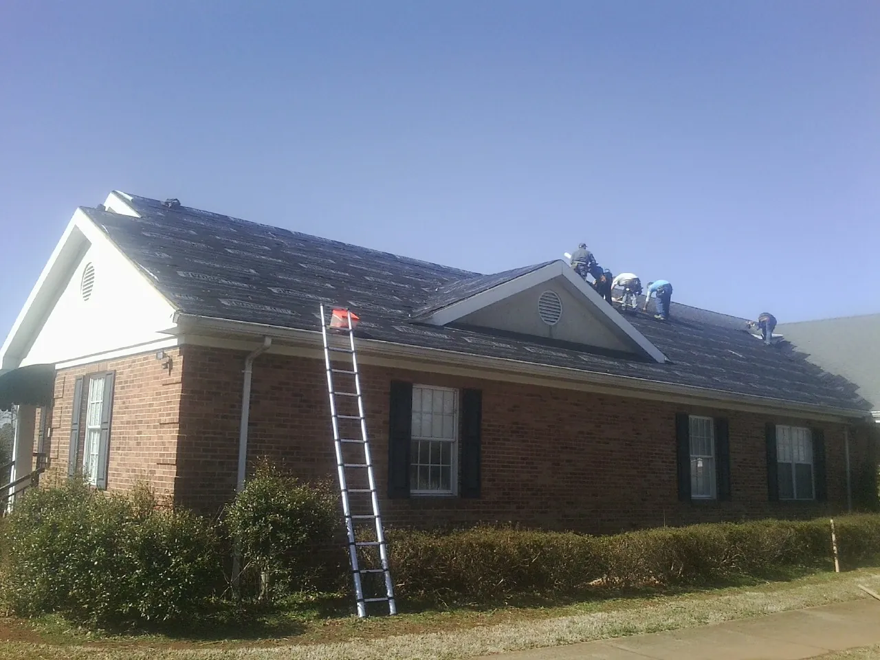 Crew tearing shingles off a brick ranch roof with a ladder set — Emergency Roof Repair work by our crew in West Blocton
