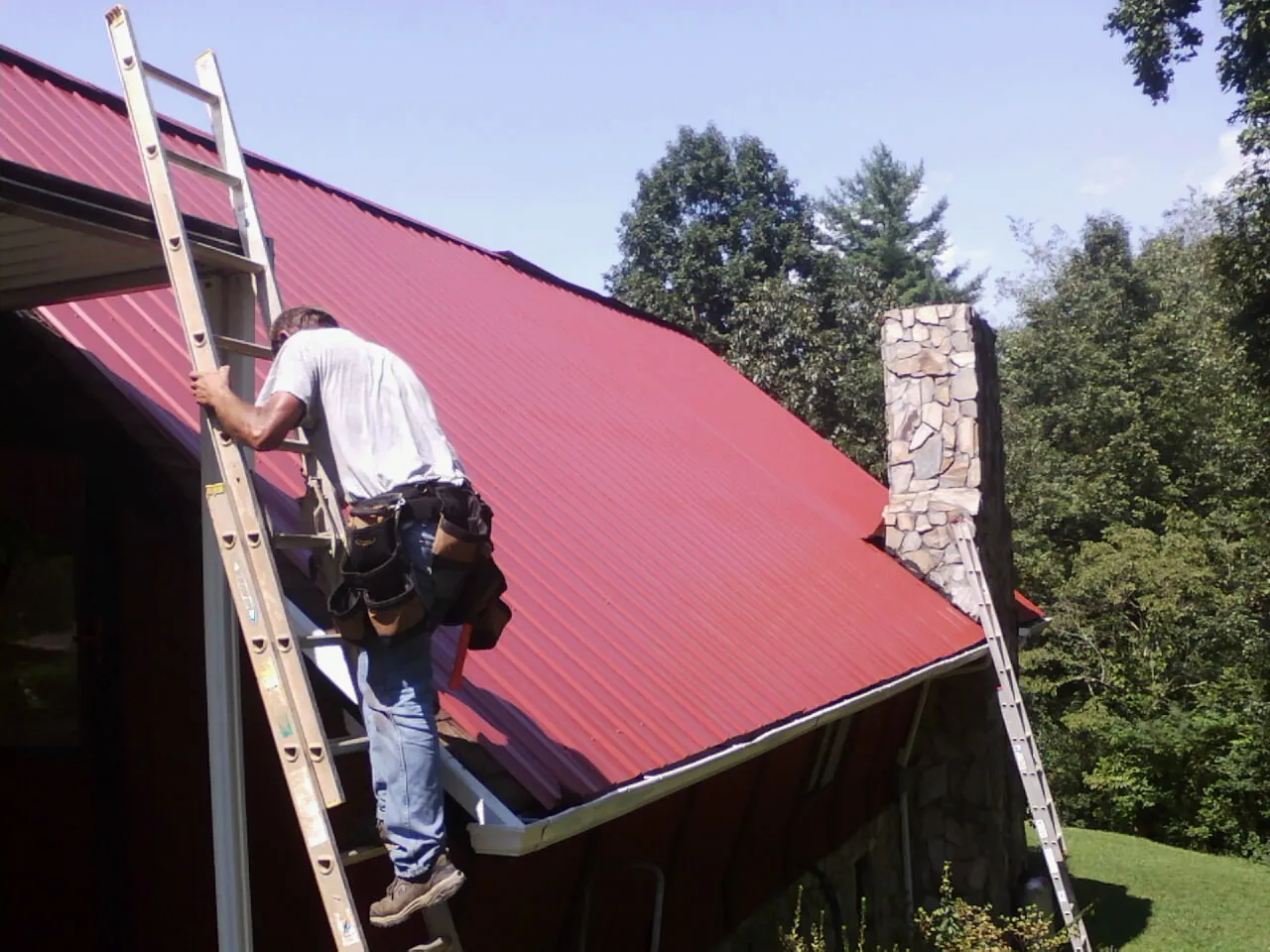 Roofer on a ladder beside a red metal roof and stone chimney — Soffit & Fascia Repair by Roofing Brothers in West Blocton